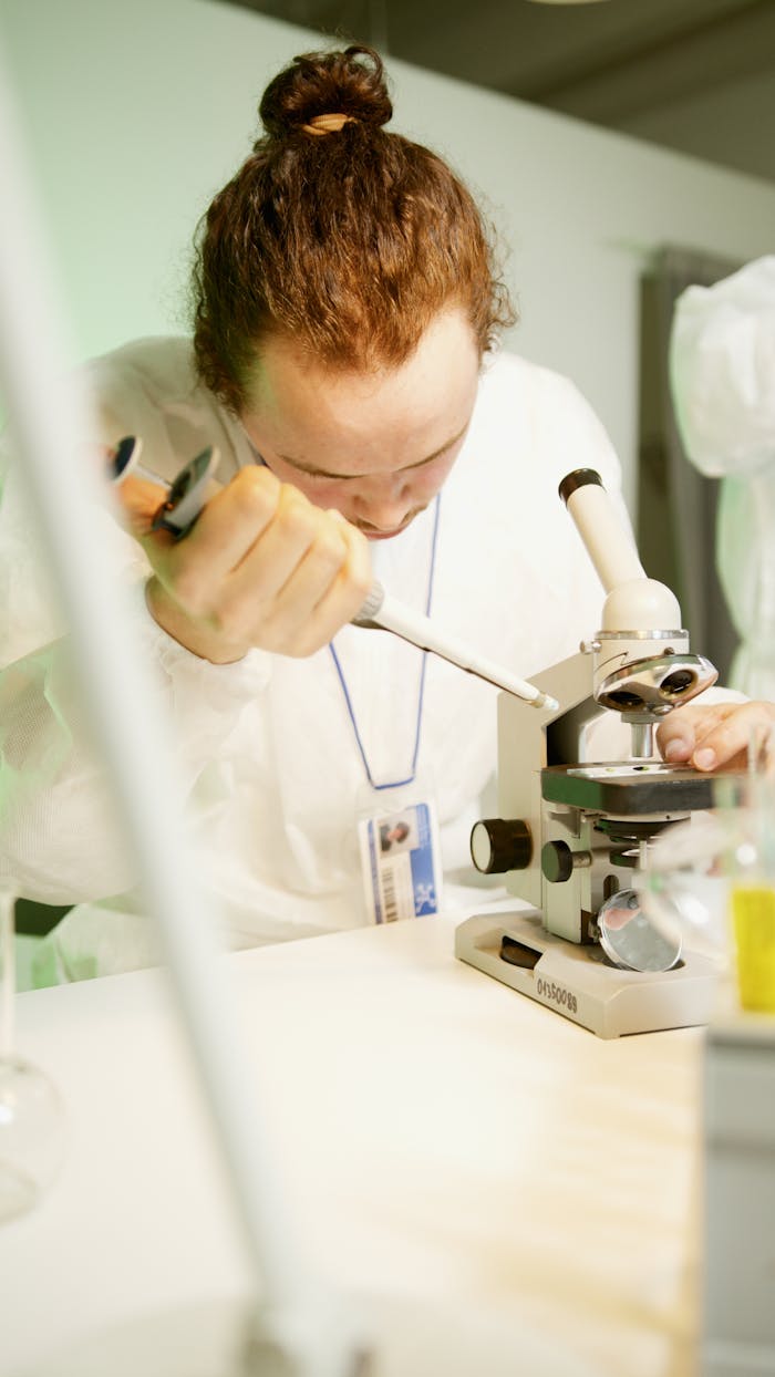 Scientist wearing a lab coat uses a microscope and pipette for research.